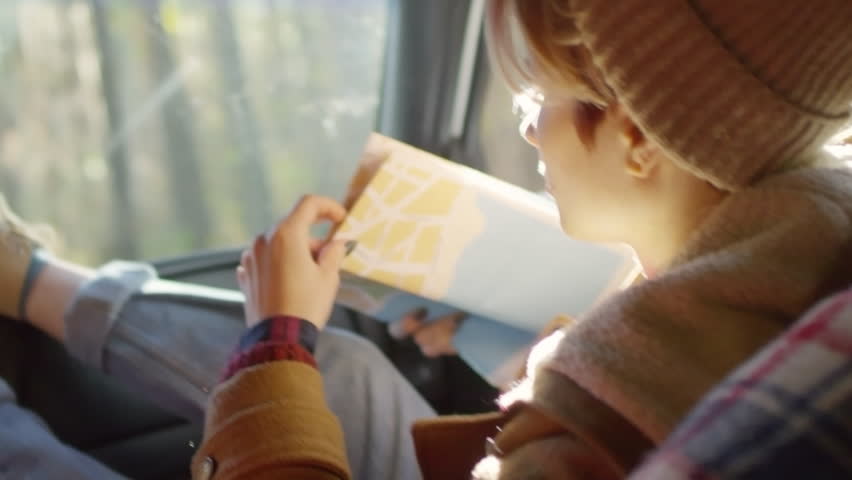 Shaky shot of young Caucasian woman using map while traveling by car and talking to unrecognizable driver