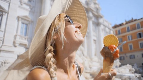 View of young woman in Rome in summer eating an Italian ice cream gelato in front of Trevi fountain at daytime enjoying capital city travel vacations - Tourist caucasian female 