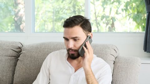 Young man sitting on sofa working on his laptop and talking on phone to customer. White male with beard, freelence concept.