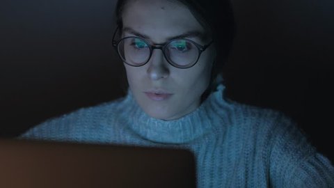 Panning shot - Portrait of a female working at the computer in the night, the reflection of the laptop screen with her glasses, woman browsing the Internet.