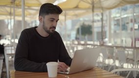 Focused young man using laptop in cafe. Handsome bearded concentrated freelancer typing on laptop computer while sitting at wooden table with coffee to go in outdoor cafe. Remote work concept