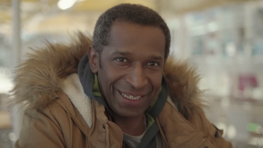 Middle aged man in winter jacket smiling at camera. Handsome cheerful African American man in warm clothes laughing and smiling at camera in outdoor cafe. Emotion concept | Shutterstock HD Video #1023567685