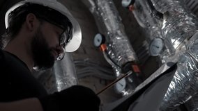 Boiler heating system inspection: man with beard, wearing in helmet makes note, checks pipes with pressure indicares. Technician (engineer) works in boiler room or ventilation room, checks the systems - Powered by Shutterstock - Get 15% off with code: PIKWIZARD15