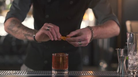 Unrecognizable bartender squeezes orange rind into a glass with a cocktail, then puts the rest into the glass close up. Bartender making alcoholic cocktail in modern bar