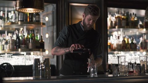 Professional bearded bartender pouring rum in the metal beaker, then in glass. Barman making cocktail in modern bar with many bottles on shelves