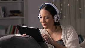 Excited woman wearing headphones finding online content on tablet sitting on a couch in the night at home
