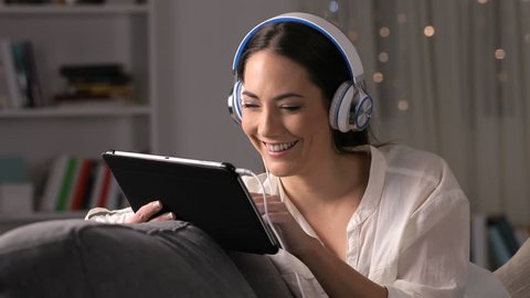 Happy girl wearing headphones watching media on tablet sitting on a couch in the night at home