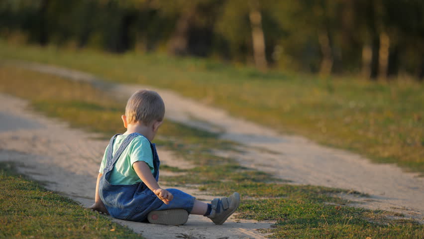 Baby child sitting on countryside road and playing with dust, toddler with back, magical; sunset light