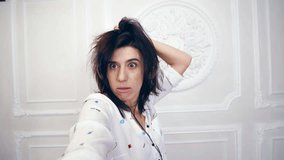 portrait of funny young woman, girl, brunette, in white shirt, in headphones, she listening to music, shoots video selfie over white wall with decorative stucco, the girl makes funny, silly faces,