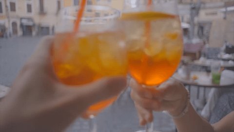 Point of view of two people cheering with glass of orange cocktail in a bar in Rome, Italy; Europe. Happy couple celebrating with aperitif drinks in Piazza having fun