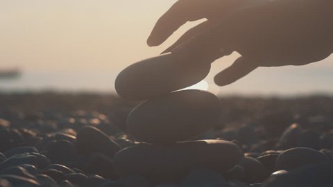 Woman hand building tower from pebbles on beach at sunset. Human hand building balanced pebbles pyramid on sea coast at sunrise. Relaxation at sea beach in evening at summer vacations