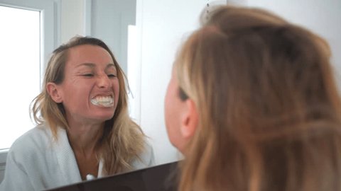 Young woman in bathroom brushing teeth in the morning looking at herself into mirror. People healthcare and medicine concept