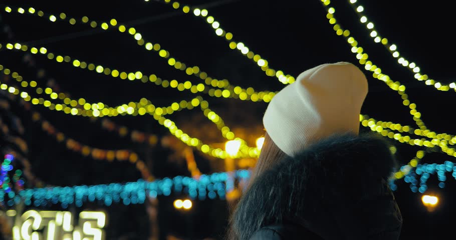 Girl in a white hat and mittens goes and smiles around the city at night. City is decorated with lights. Circular motion of the camera.
