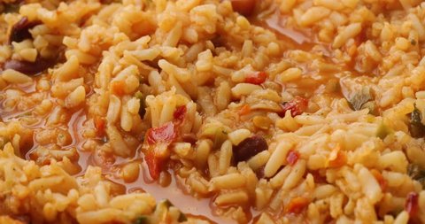 Close video of Mexican rice and beans being stirred with a spoon while cooking in a skillet and taking a spoonful at the end illuminated with natural lighting.