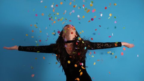 SLOW MOTION: Young woman having fun with confetti spreading arms and smiling. Blue studio background.