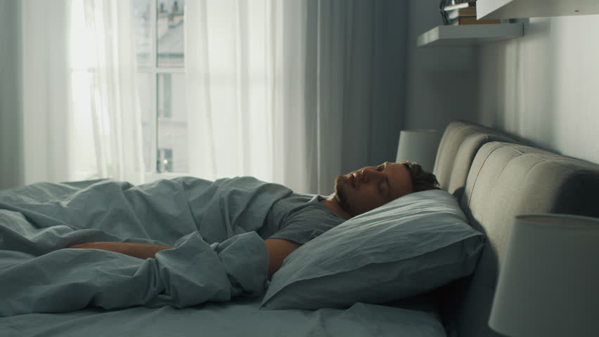 Handsome Fair Boy Sleeping Cozily in Bed, Slowly Wakes up, Gets out of Bed and Stretches Lazily. Young and Fit Caucasian Man Greeting New Day. Early Morning Sun Shines Through the Window. Royalty-Free Stock Footage #1023631615