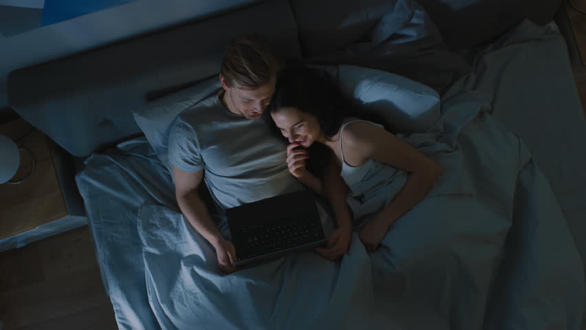 Cute Young Couple in Bed at Night Using Laptop, Watching Series, Reading Social Media, Browsing Through Internet, Talking and Having Fun. Top View Camera Shot - Powered by Shutterstock - Get 15% off with code: PIKWIZARD15