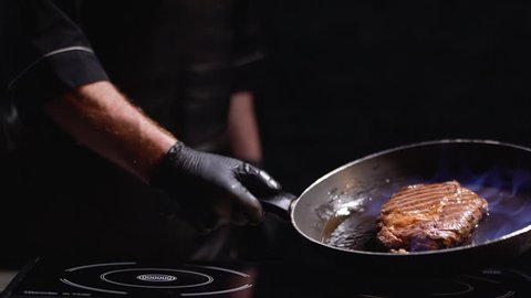 Male hand in black rubber gloves holding a frying pan with a grilling steak. The cook turns the pan on the fire in different directions. Slow motion.