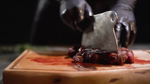 Male hand in black rubber gloves chops meat lying on a cutting board with big knife close up.