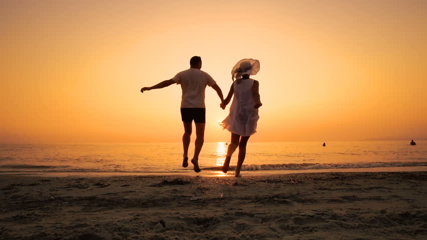 Romantic couple running on beach at sea sunset holding hands and jumping in slow motion. Man takes woman in air and turning her around. Concept of love and happiness