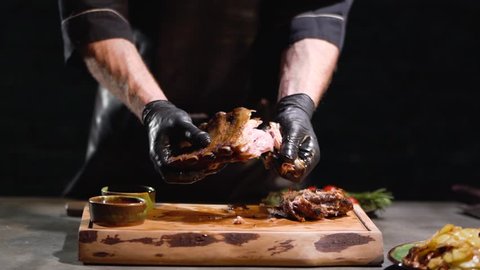 Male hands in black rubber gloves tears apart juicy roasted ribs close up. Slow motion.