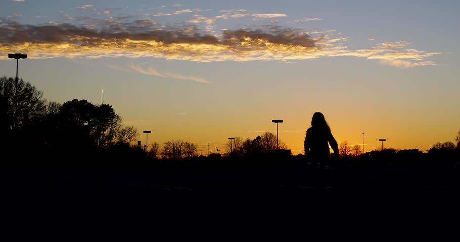 Silhouette of a young girl with long hair dancing, jumping and running in a parking lot with a beautiful blue and orange sunset in the background with some clouds, trees and lights in the scene. 4K