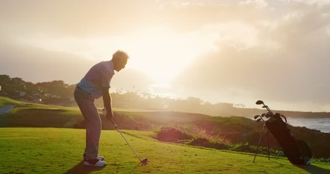 Handsome older golfer swinging and hitting golf ball on beautiful course at sunset by the ocean, slow motion