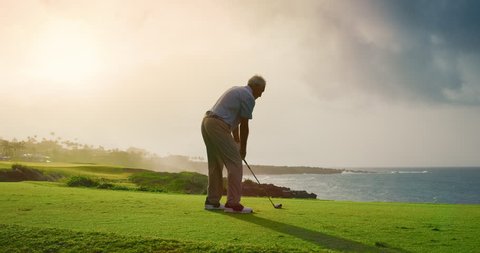 Handsome older golfer swinging and hitting golf ball on beautiful course with ocean at sunset, slow motion
