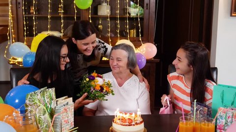 Senior mother with daughter and granddaughters with flowers and present at birthday party. Celebrating life and love concept between generations. Mothers day concept. Slow motion 4k hand held movement