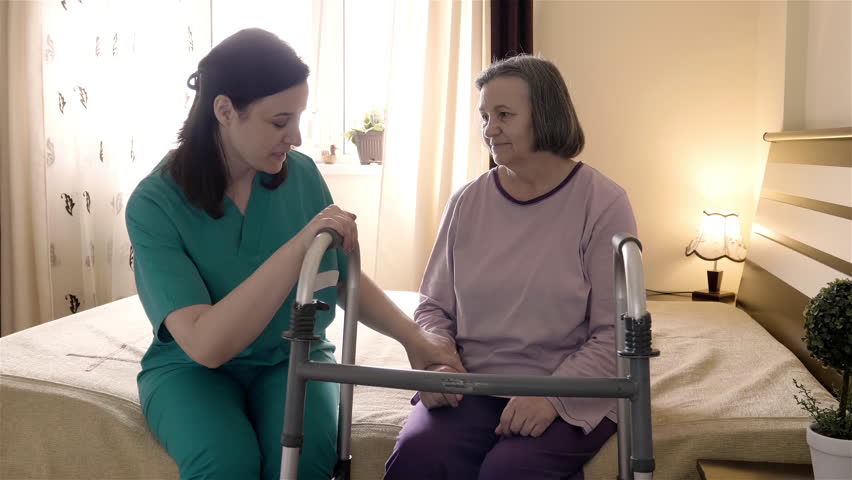 Friendly caregiver helping senior woman getting up from bed and walk with a walker. Home or hospice nursing and assistance concept. Slow motion 4K | Shutterstock HD Video #1023671869