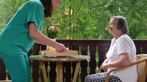 Pan of smiling young nurse brings breakfast to happy senior woman at nursing home. Slow motion 4k