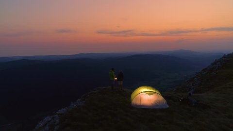 DRONE: Flying above a young couple talking by the lantern on a calm summer evening in the picturesque Alps. Carefree man and woman enjoying a romantic evening in the breathtaking serene mountains.