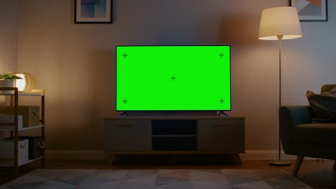 Zoom In Shot of a TV with Horizontal Green Screen Mock Up. Cozy Evening Living Room with a Chair and Lamps Turned On at Home.