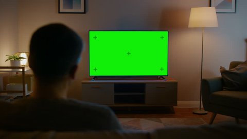 Young Man in Glasses is Sitting on a Sofa and Watching TV with Horizontal Green Screen Mock Up. It's Evening and Room at Home Has Working Lamps.