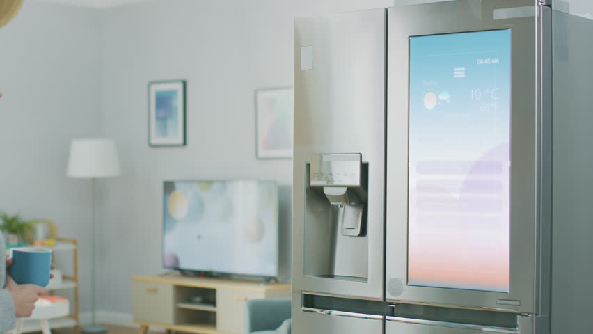 Beautiful Young Girl Walks Over to a Refrigerator While Drinking Her Morning Coffee. She is Checking the Weather Forecast and a To Do List on a Smart Fridge at Home. Kitchen is Bright and Cozy. Royalty-Free Stock Footage #1023697405