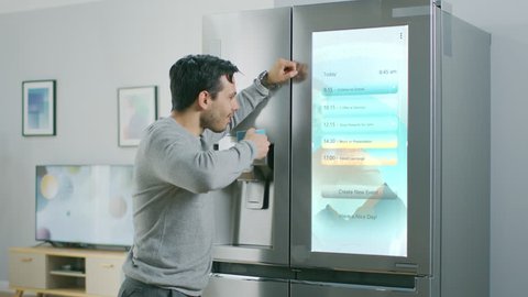 Handsome Young Man Walks Over to a Refrigerator While Drinking His Morning Coffee. He is Checking a To Do List on a Smart Fridge at Home. Kitchen is Bright and Cozy.