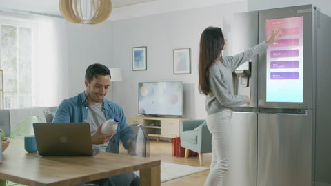 Beautiful Young Woman Opens the Fridge and Gives a Milk Bottle to Her Boyfriend. Then She Checks the Futuristic Digital To-Do List on the Smart Fridge Door.