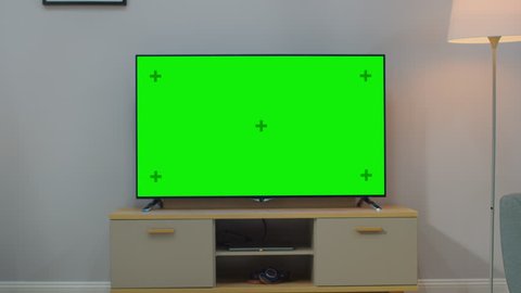 Zoom Out Shot of a TV with Horizontal Green Screen Mock Up. Cozy Living Room at Day Time with a Chair and Lamps Turned On at Home.