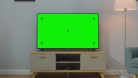 Zoom In Shot of a TV with Horizontal Green Screen Mock Up. Cozy Living Room at Day Time with a Chair and Lamps Turned On at Home.