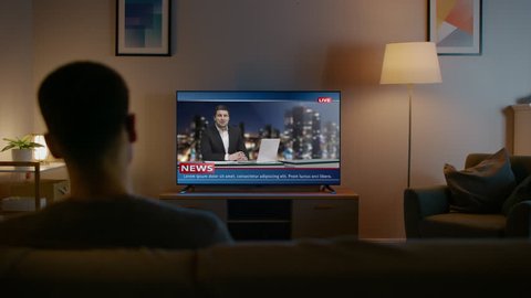 Young Man in Glasses is Sitting on a Sofa and Watching TV with Live News. It's Evening and Room at Home Has Working Lamps.