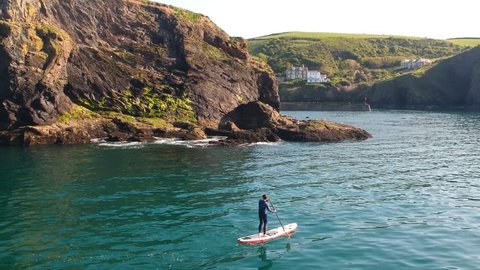 Paddleboarder moving calmly through blue waters below cliffs in Cornwall, England. The shot slowly pans to the right, showing more of the countryside beyond the cliffs