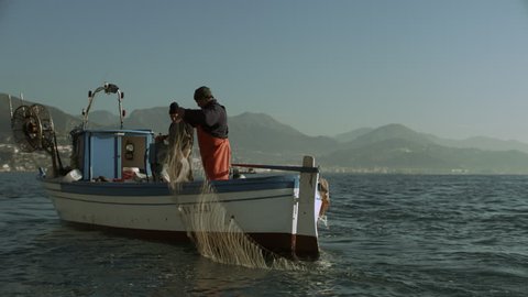 Two busy fishermen sailing on their boat across the sea with view of the Amalfi Coast in the background, on a bright sunny day. Wide shot on 8k helium RED camera.
