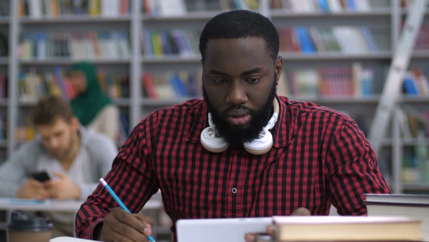 Trendy african american male student in modern headphones with digital tablet pc making distance online learning in academic library. Dark-skinned student e-learning through video conference app.