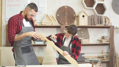 Little male woodworker creating his dream and his Dad is helping him. Close-up of young male carpenter examines with his son smoothness of DIY wooden sword to work with wood in his workshop