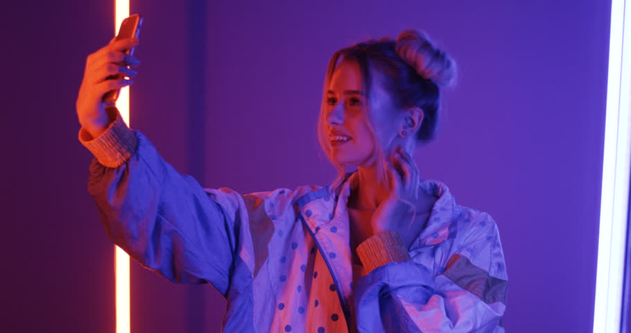 Cute blonde young woman in stylish outlook taking selfie pictures on the smartphone camera and posing in the violet neon light.