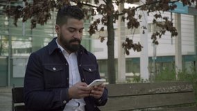 Focused bearded man messaging via smartphone on street. Concentrated handsome man sitting on wooden bench and using mobile phone outdoor. Technology concept