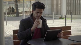 Man drinking from paper cup and using digital tablet. Serious handsome young man drinking coffee to go and working with tablet pc in outdoor cafe. Remote work concept