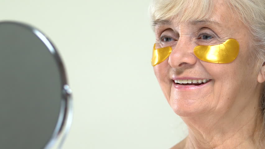 Senior female with anti-aging hydrogel eye patches on face looking in mirror