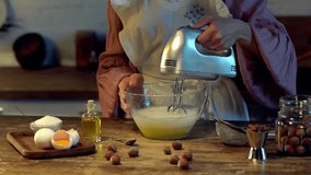 female cook whipping eggs in glass bowl with mixer at wooden table with raw ingredients