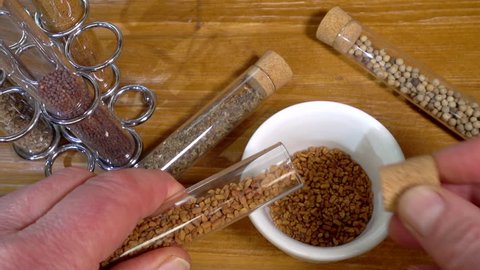 Slow motion close POV overhead shot of a man's hands pouring fenugreek seeds into a small dish then replacing the cork, from one of a range of glass tubes containing spices, on a pine kitchen worktop.